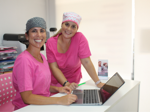 Trato cercano y comunicación clara Dos profesionales de Presencia Dental, con bata rosa, explican un plan de tratamiento en un portátil y sonríen a cámara